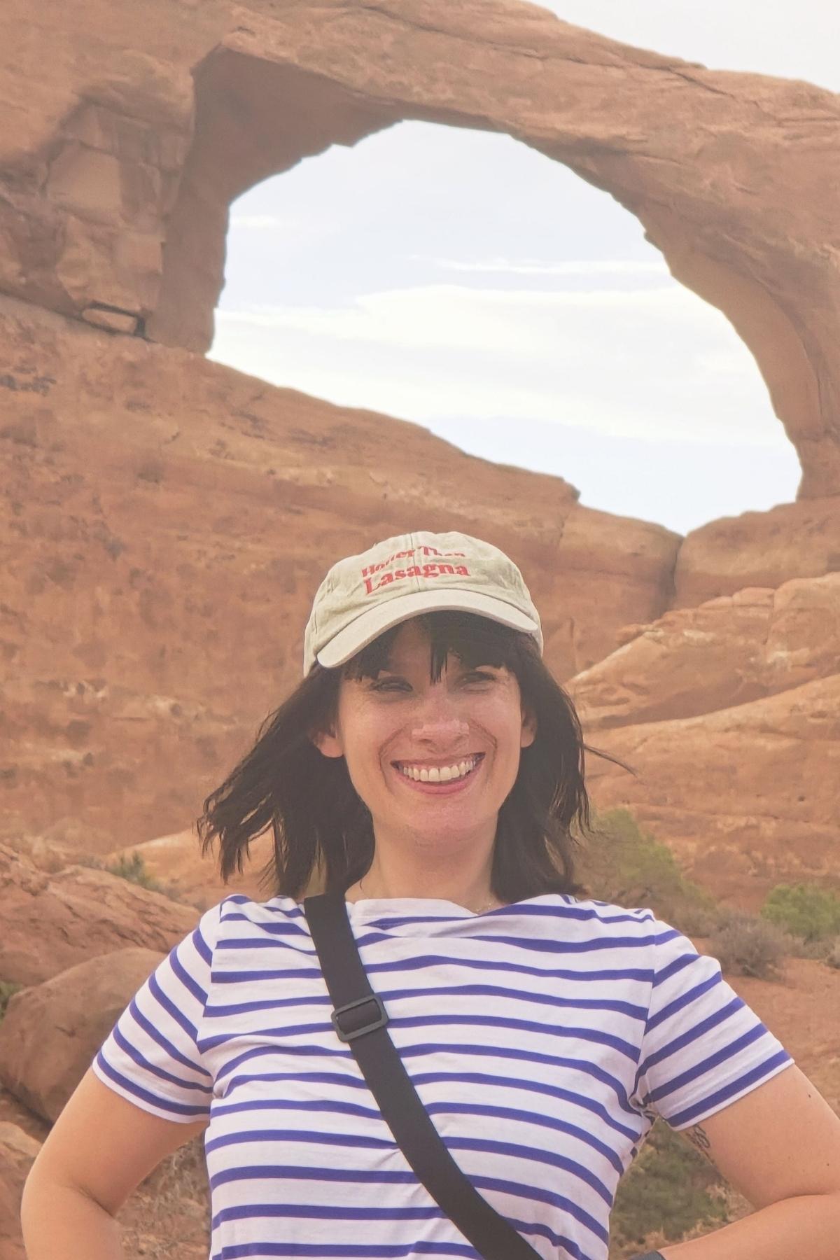 Una donna sorridente con una camicia a righe e un berretto beige è in piedi davanti a un arco di roccia naturale in un paesaggio desertico. Il cielo è leggermente coperto.