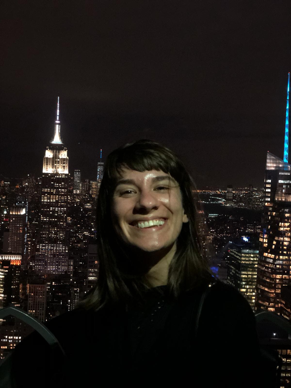 Alice Zilio travel designer sorridente di fronte a un paesaggio urbano notturno, con l'Empire State Building illuminato e le luci della città sullo sfondo, che trasmettono un'atmosfera gioiosa. Si trova a New York, in cima al top of the Rock.