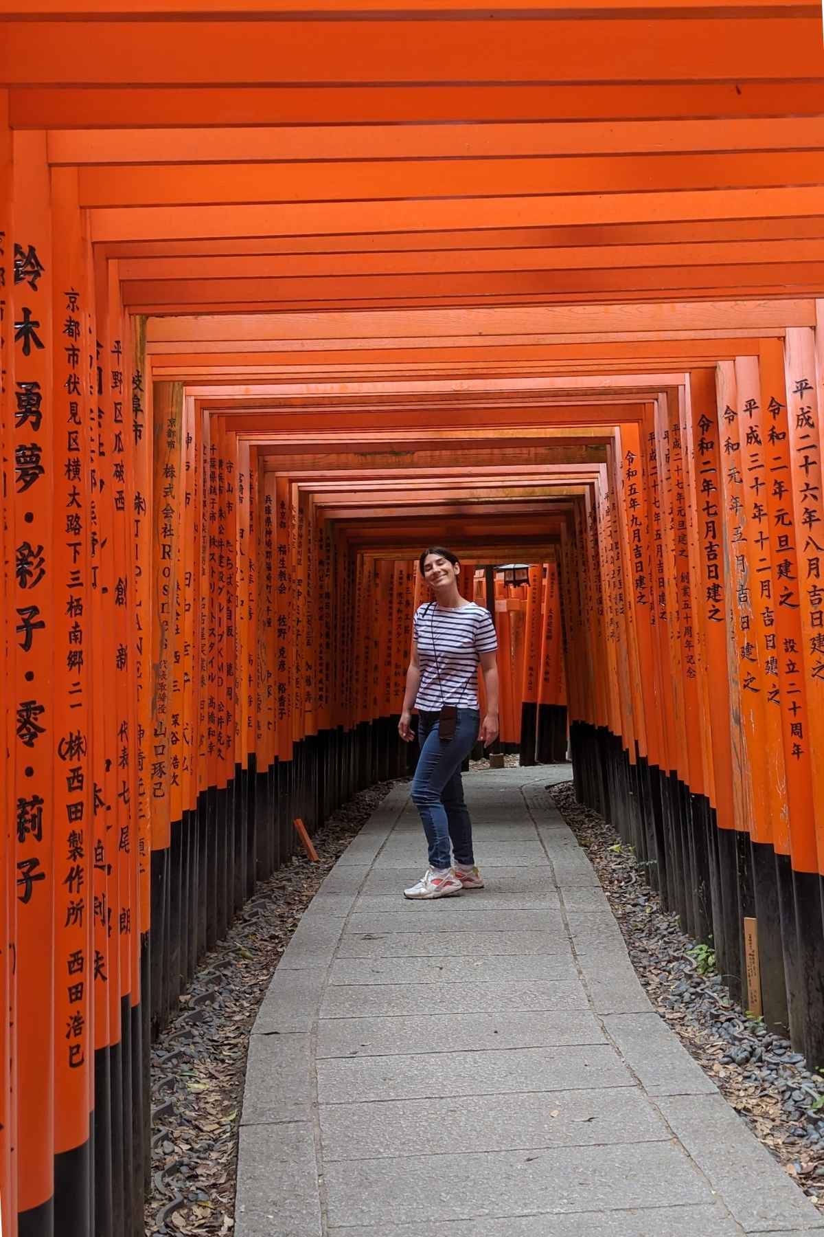 Alice Zilio travel designer attraversa gli iconici torii arancioni del santuario di Fushimi Inari in Giappone. La scena è serena, con caratteri giapponesi sui cancelli.