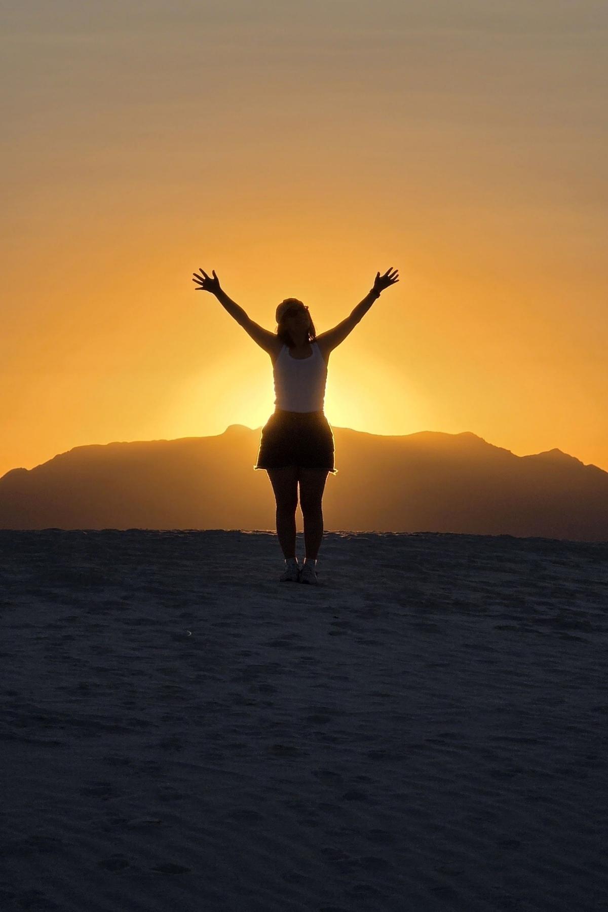 alice-zilio-travel-designer-tramonto-a-white-sands-new-mexico Alice Zilio travel designer con le braccia alzate, in piedi nel parco nazionale americano di white sands in new mexico, al tramonto. Il sole tramonta dietro le dune lontane, proiettando un caldo bagliore dorato.