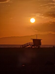 los-angeles-venice-beach-tramonto Tramonto su una spiaggia con la sagoma di una torretta di salvataggio in primo piano. Il cielo è arancione e calmo, creando un'atmosfera di pace. La spiaggia è venice beach